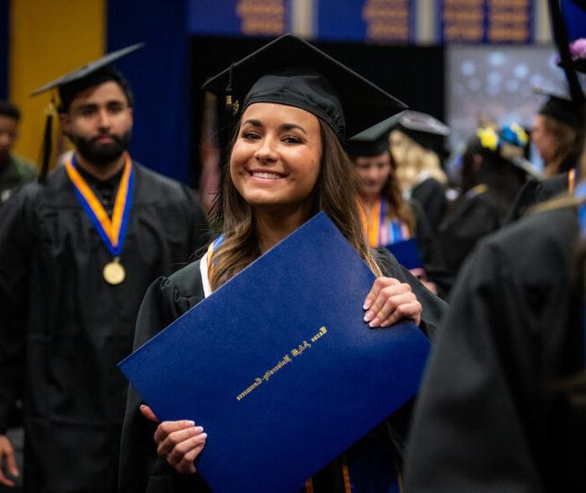 Graduate holds diploma in both hands and smiles at the camera, wearing graduation cap and gown.